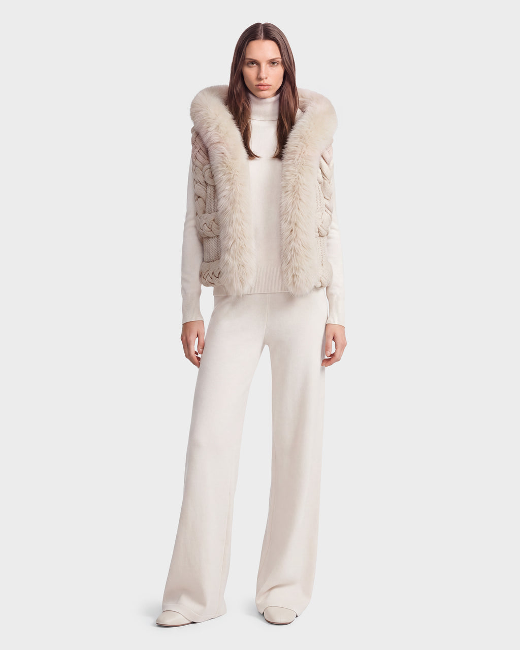 A woman stands against a plain light background wearing the Rudsak CICELY - 8526686 Vanilla—a cream turtleneck sweater featuring a sleeveless knit vest with a faux fur-trimmed hood—paired with wide-legged pants, creating an ensemble perfect for winter layering.