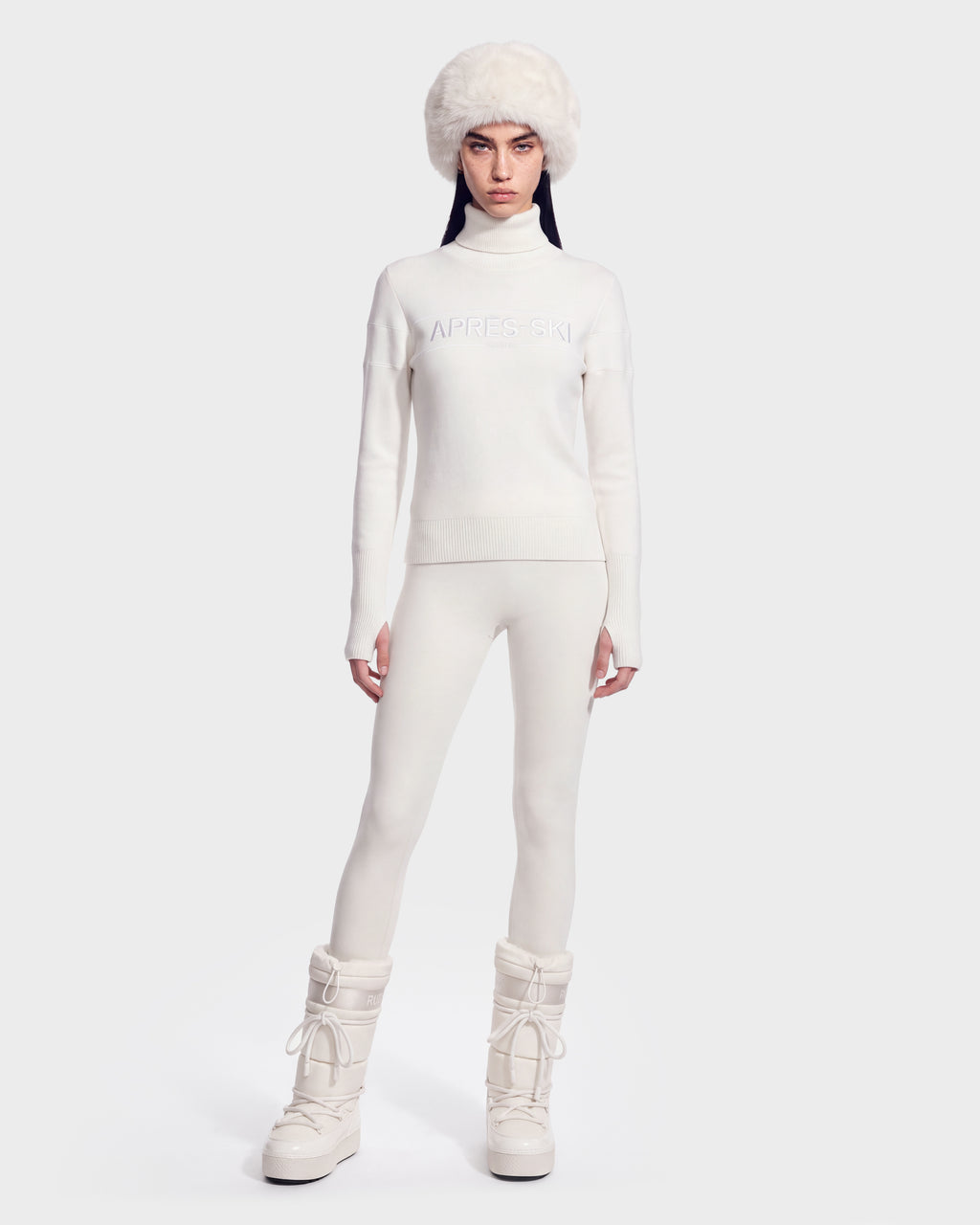 A woman wearing the ERIKA - 8525705 Iceland apres-ski turtleneck sweater by Rudsak pairs her all-white winter look—including a fuzzy white hat, white leggings, and snow boots—while standing against a plain light background.