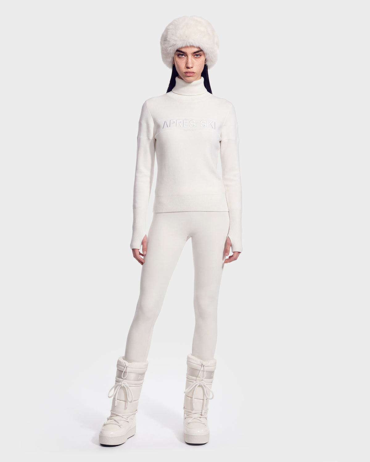 A woman wearing the ERIKA - 8525705 Iceland apres-ski turtleneck sweater by Rudsak pairs her all-white winter look—including a fuzzy white hat, white leggings, and snow boots—while standing against a plain light background.