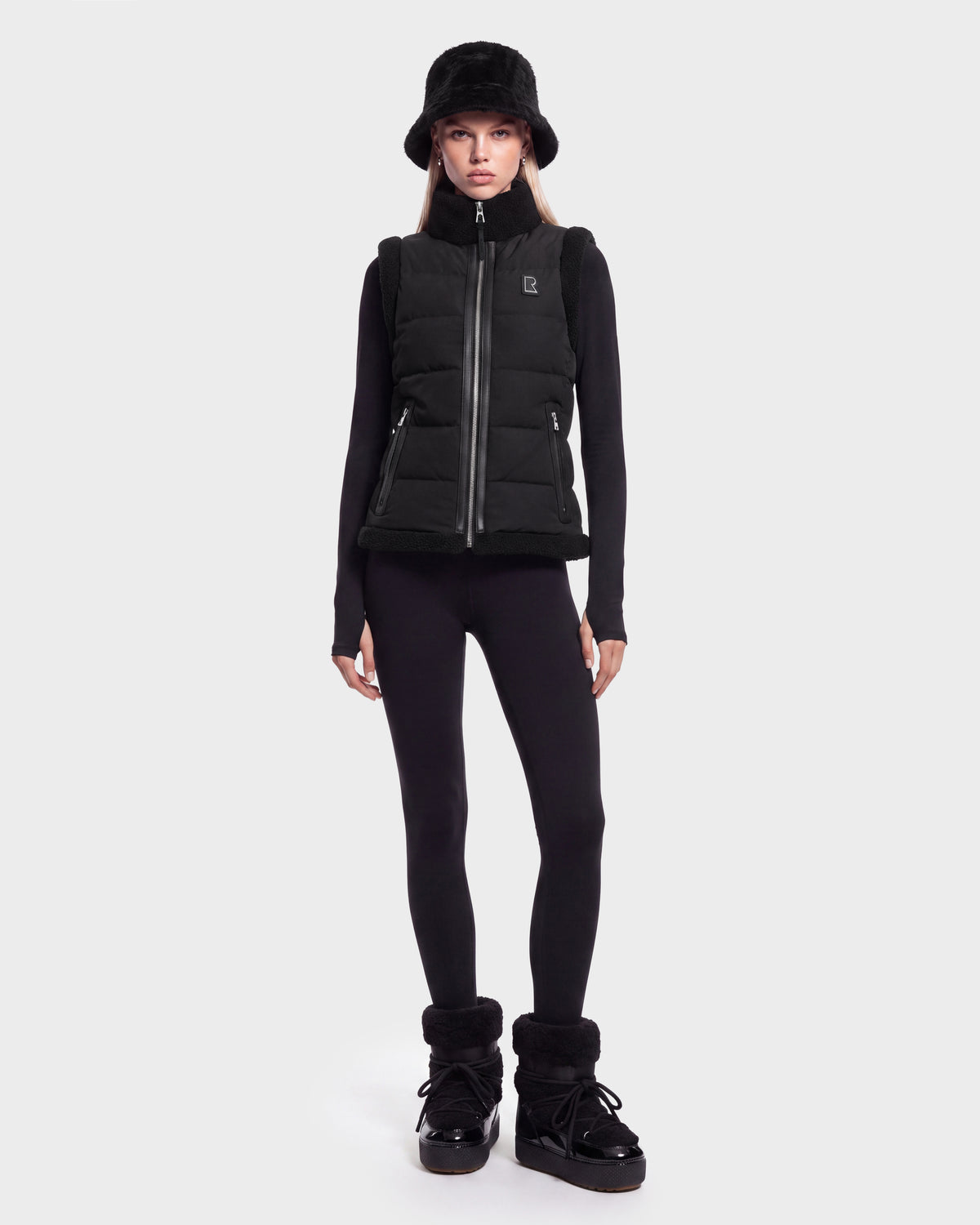 A woman stands facing forward against a plain white background, wearing the Rudsak HALI - 8126771 Black puffer vest filled with RDS-certified down for lightweight warmth, paired with a black bucket hat, long-sleeve black top, black leggings, and black boots.