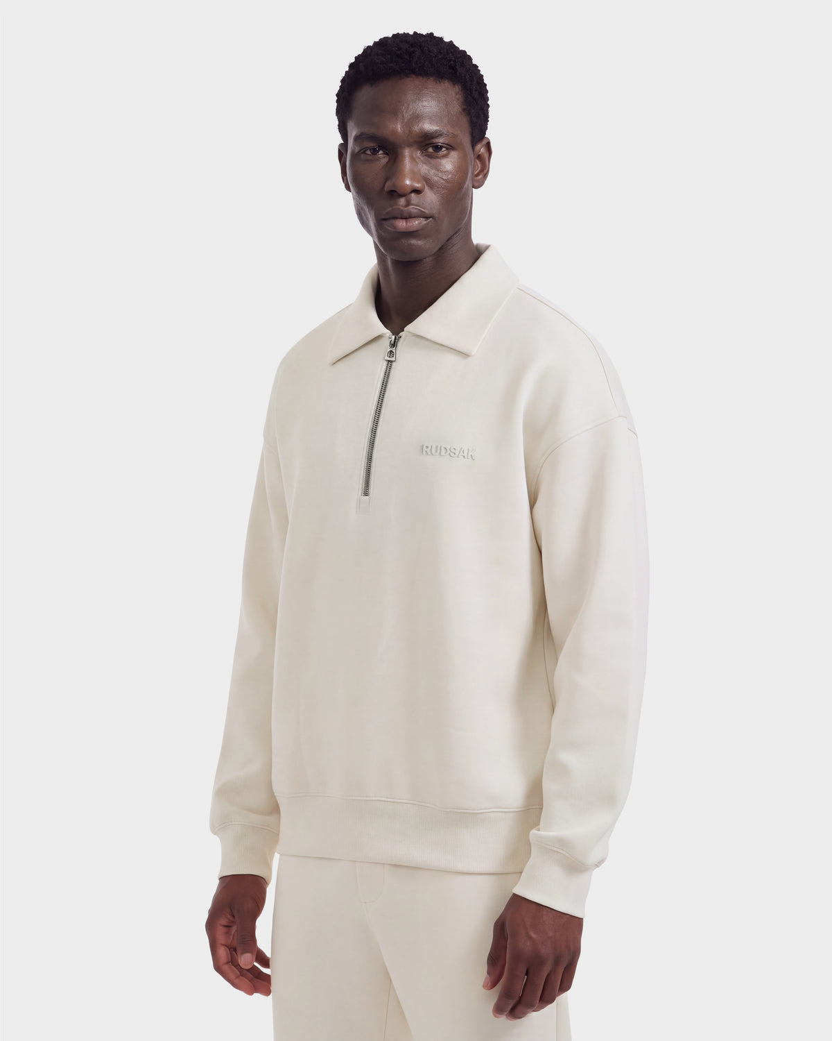 A man wears a cream-colored, long-sleeve IDRIS - 6526500 Vanilla quarter-zip polo sweater with a relaxed silhouette and collared neckline.