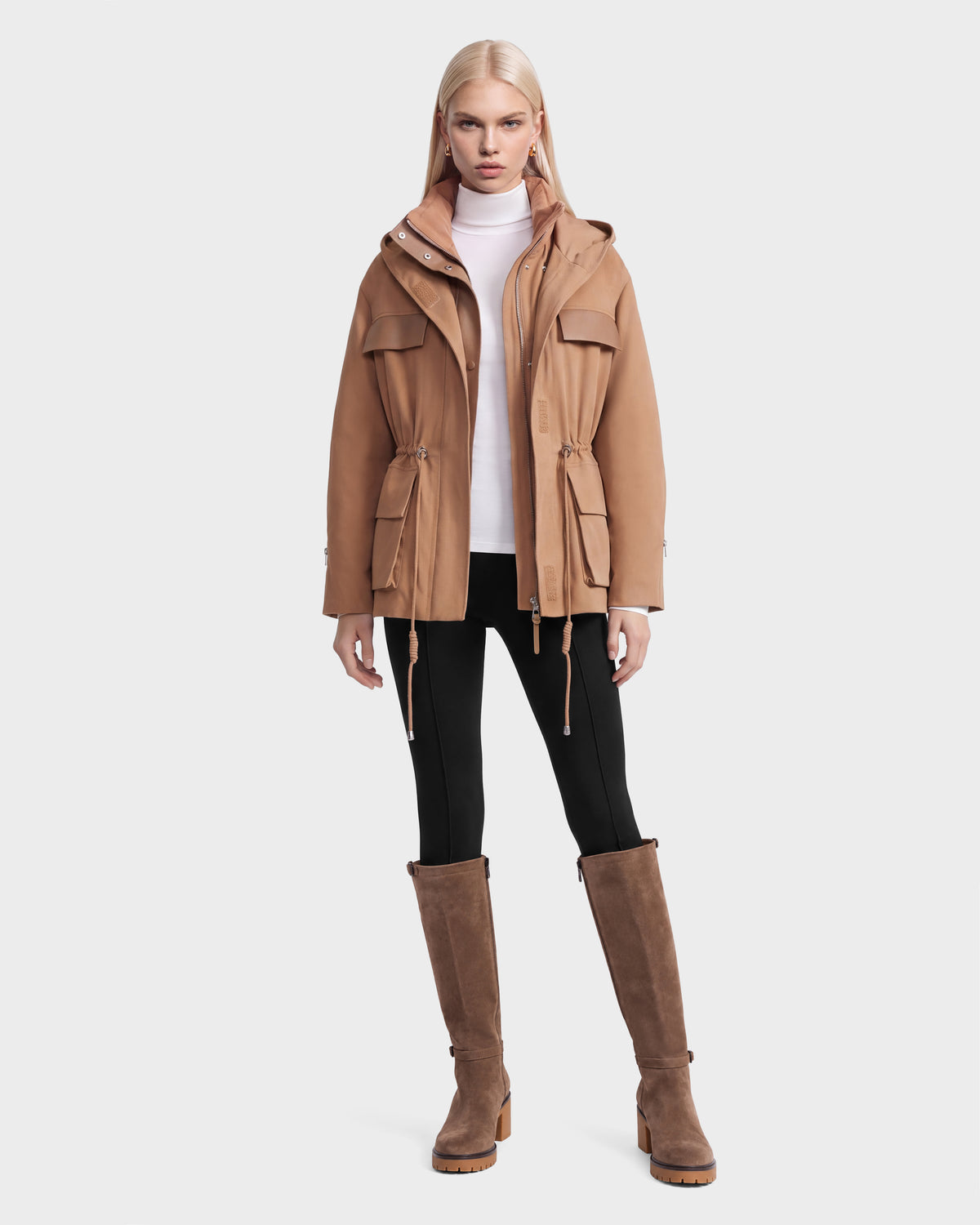A model wears a camel-colored ROSALINDA - 8126523 hooded jacket with a drawstring waist, a white turtleneck, black pants, and knee-high brown boots.