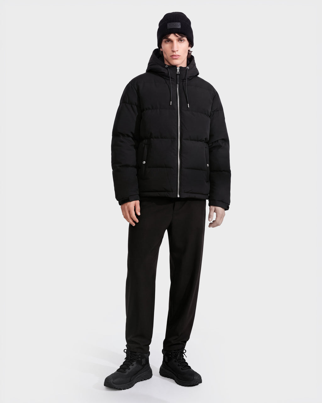 A person stands facing forward against a plain white background, wearing the Rudsak STERLING EL - 6126759 Black puffer jacket, paired with a black beanie, black pants, and black shoes.
