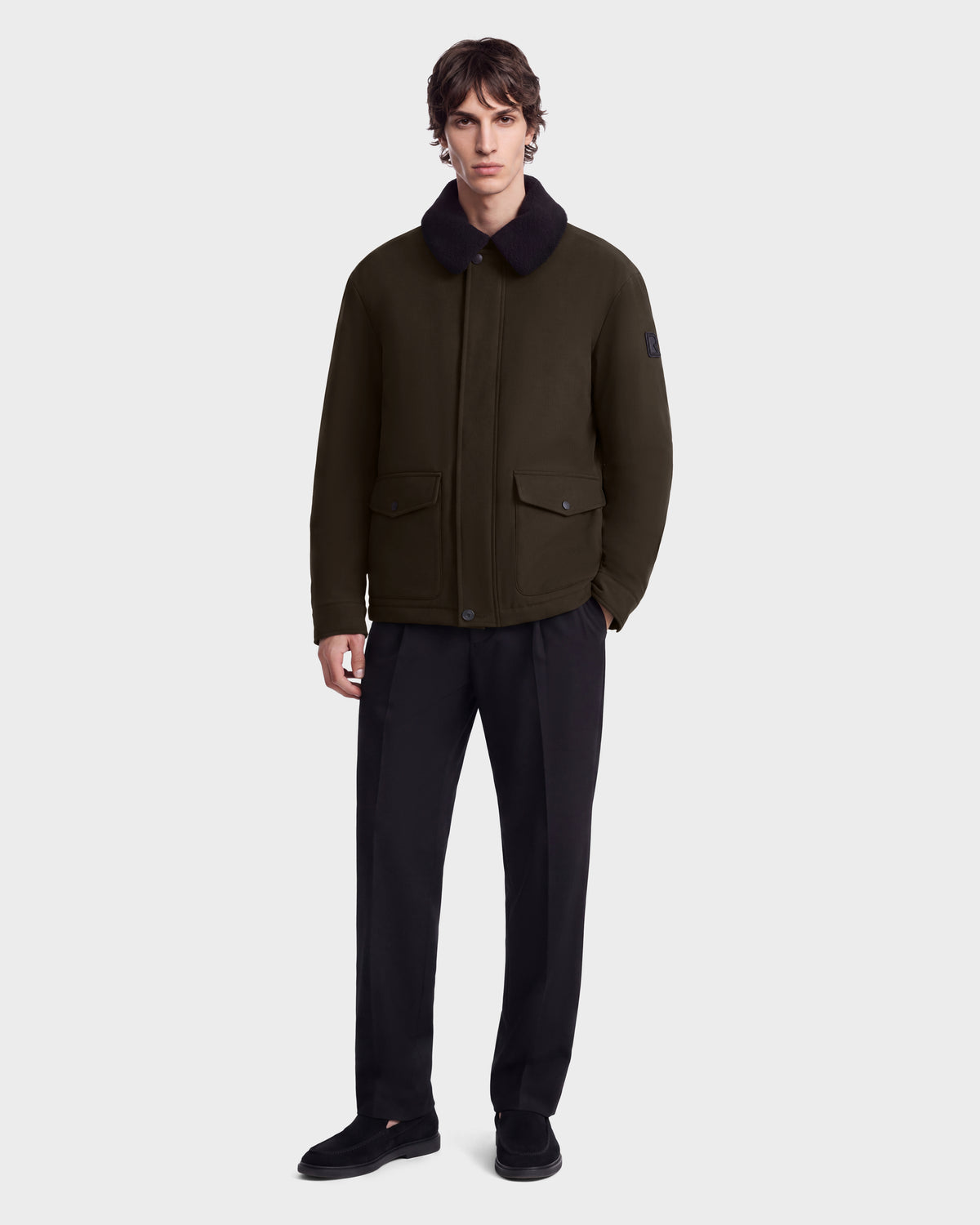 A person wearing the Rudsak ANTOINE - 6126506 Olive utility jacket with a dark faux-fur collar, dark trousers, and black loafers.