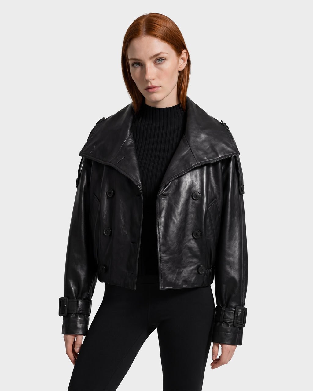 A person with straight red hair wears the Rudsak JANELLE - 8126936 Black leather bomber jacket, featuring wide lapels, buckle cuffs, and a modern silhouette, layered over a black ribbed sweater and black pants while standing against a plain light background.