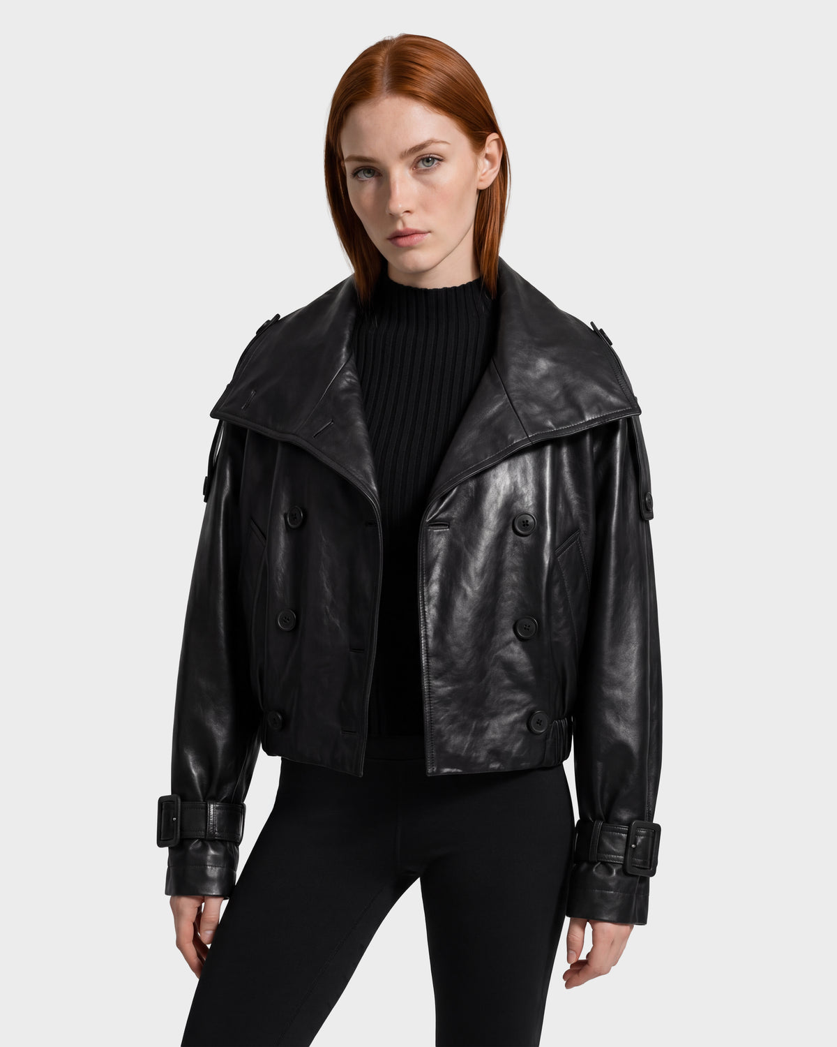 A person with straight red hair wears the Rudsak JANELLE - 8126936 Black leather bomber jacket, featuring wide lapels, buckle cuffs, and a modern silhouette, layered over a black ribbed sweater and black pants while standing against a plain light background.