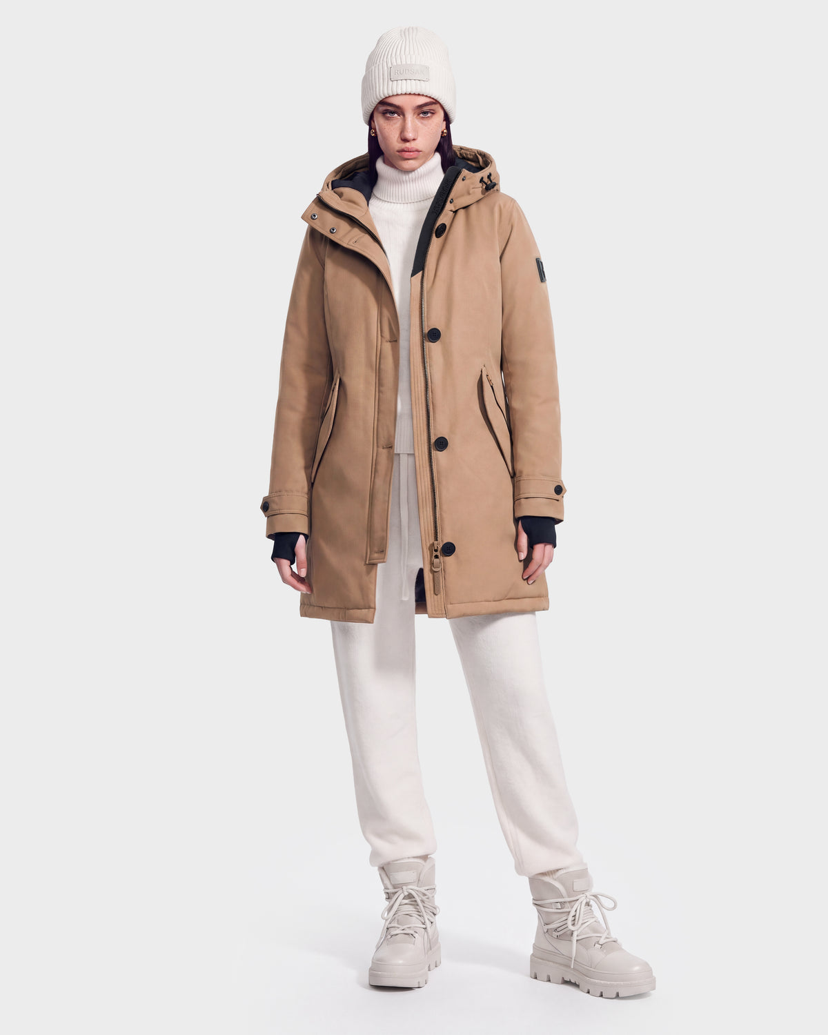 A person wearing the Rudsak KEYSHIA - 8126517 Camel parka, a white knit beanie, turtleneck sweater, cream jogger pants, and beige boots stands against a plain white background.