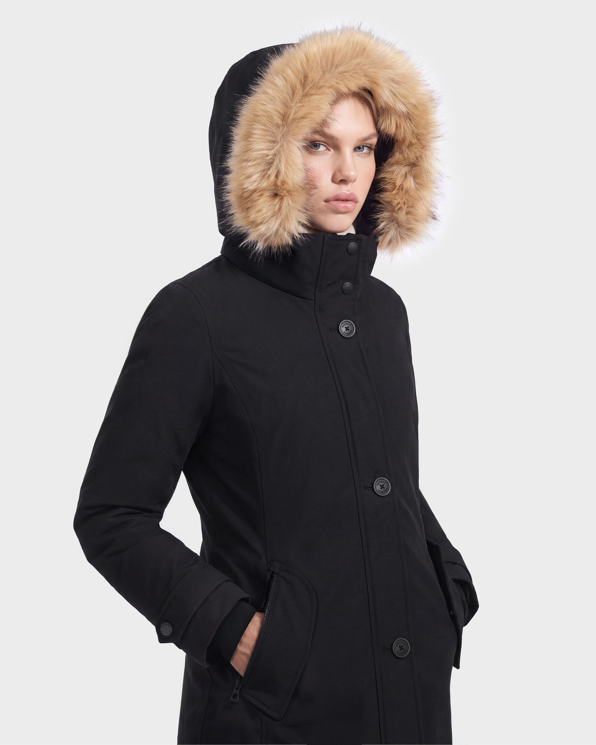 A person wears the KEYSHIA - 8126517 Black by Rudsak, a women’s down parka with a light brown faux fur-lined hood and hand pockets, standing against a simple light background—perfect for cold weather.