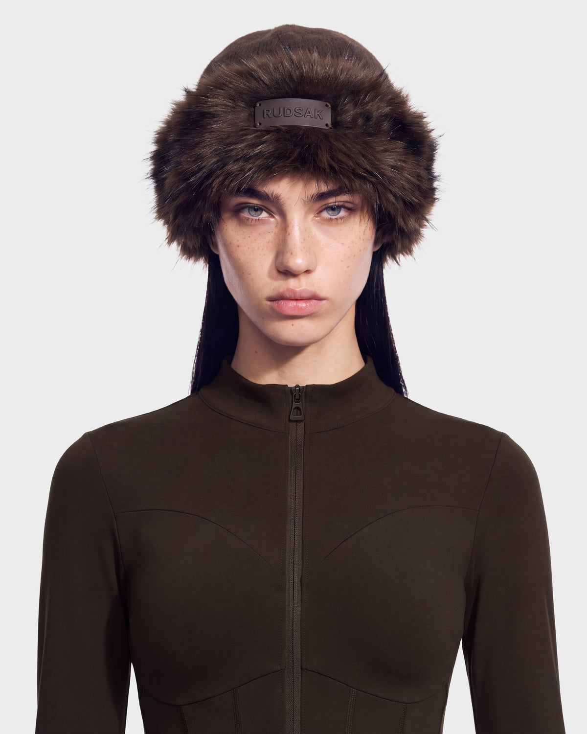 A person wearing a dark brown RUDSAK LORETTA W - 8426510 Espresso faux fur ushanka hat with a small logo patch on the front and a matching long-sleeved, zip-up top.