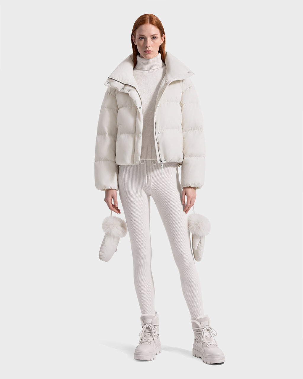 A person wears a cream-colored ALONDRA - 8126992 Vanilla cropped puffer jacket, matching turtleneck sweater, leggings, and boots, holding a pair of fur-trimmed mittens.