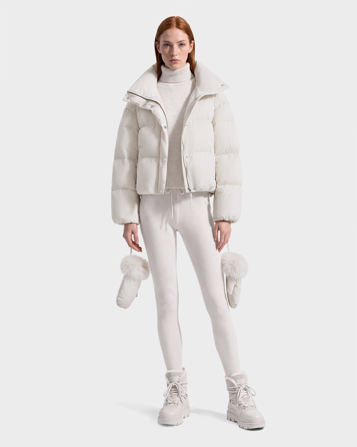 A person wears a cream-colored ALONDRA - 8126992 Vanilla cropped puffer jacket, matching turtleneck sweater, leggings, and boots, holding a pair of fur-trimmed mittens.
