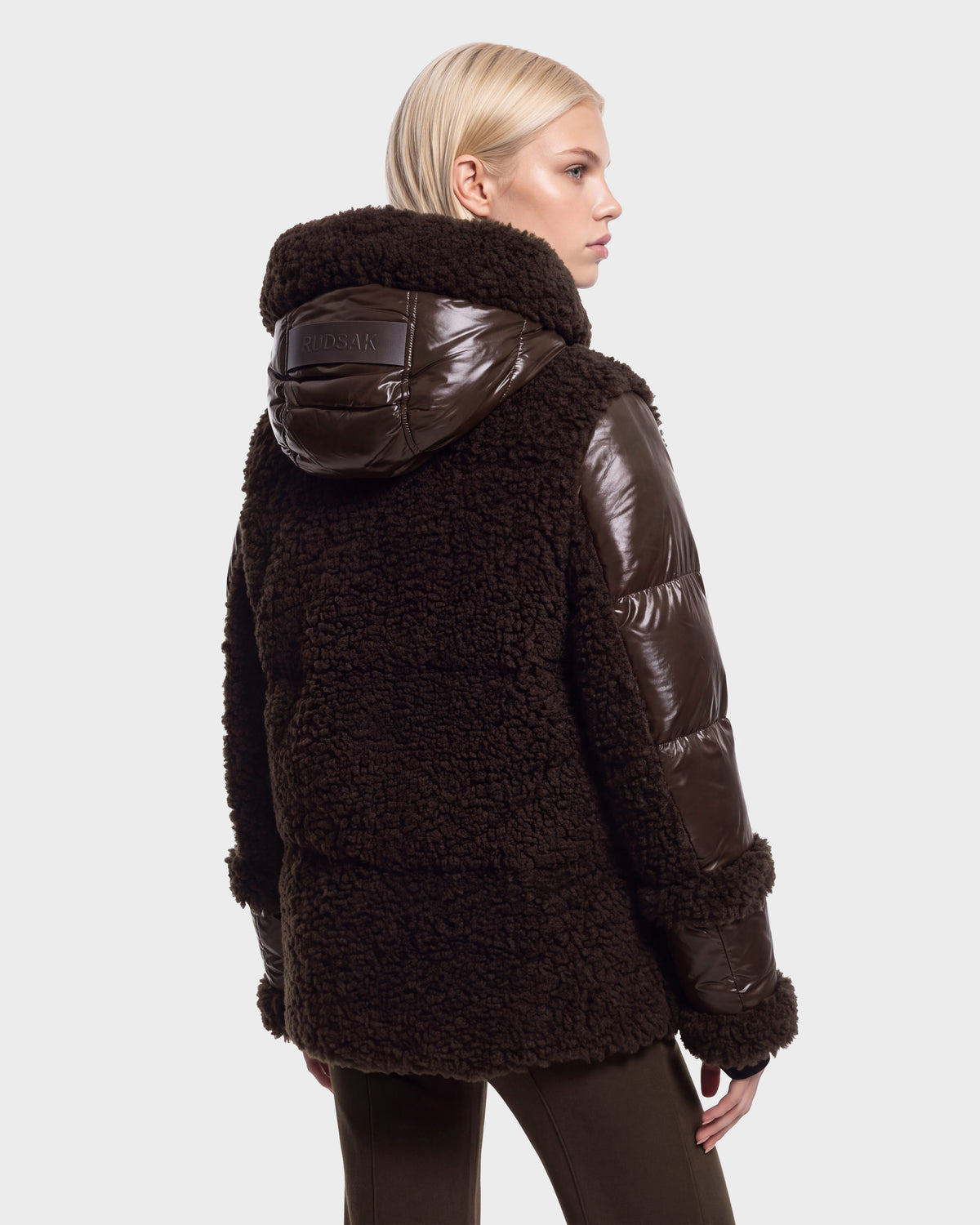 A person with short blonde hair is seen from the back against a plain white background, wearing the Rudsak MARISSA - 8126728 Espresso: a dark brown, hooded shearling and puffer jacket—perfect winter outerwear.