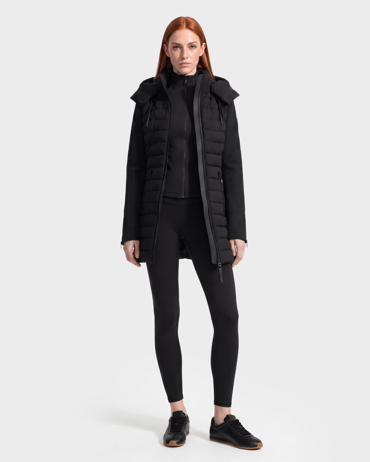 A woman with long red hair stands facing forward, wearing the MERINA - 8125056 Black quilted down jacket by Rudsak layered over a black zip-up top, paired with black leggings and black sneakers against a plain light gray background.