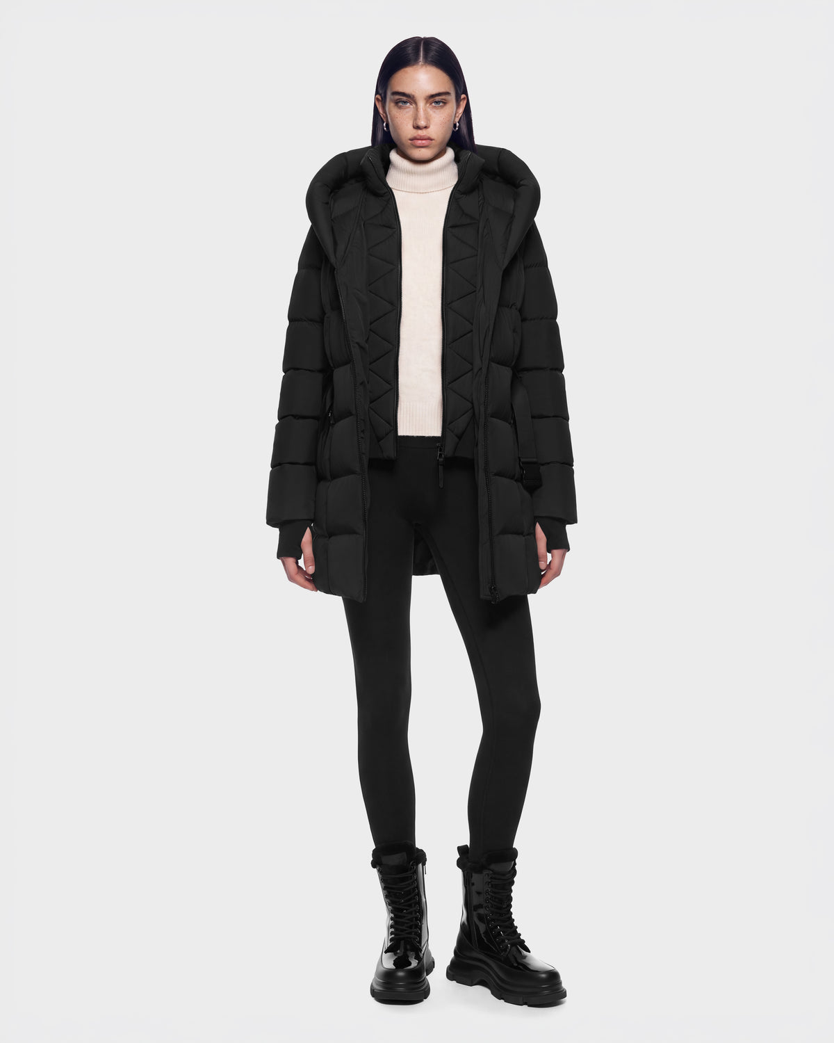 A person wearing a RUDSAK TAYLOR M - 8125965 Black down coat with a large hood, a cream turtleneck sweater, black leggings, and black lug-sole boots.