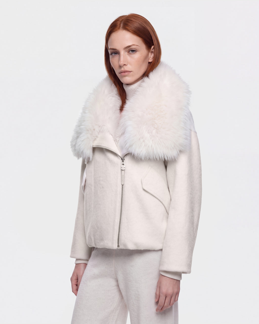 A woman with straight red hair wears the Rudsak ZARIA - 8126999 Vanilla white winter puffer jacket, which features a large, fluffy faux-fur collar, paired with white pants, standing against a plain white background.