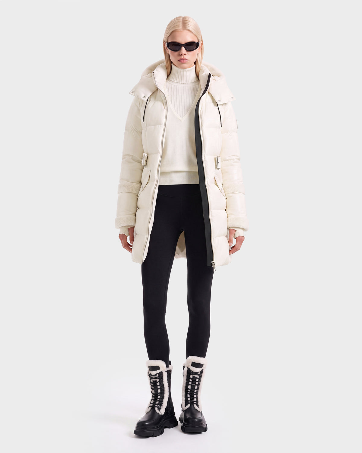 A person wearing a Rudsak AUDREY L - 8124585 Vanilla knee-length down puffer jacket, white turtleneck sweater, black leggings, and lace-up boots with fur trim.