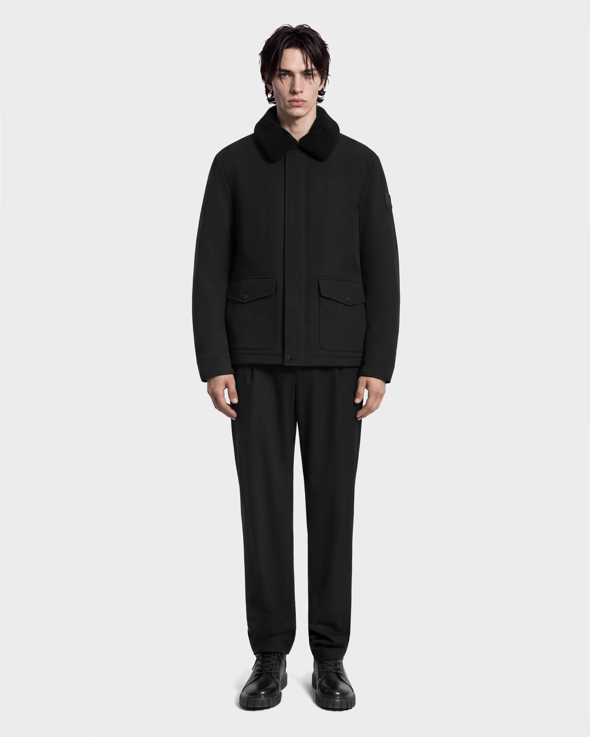 A man wearing a Rudsak ANTOINE - 6126506 Black utility jacket with a faux-fur collar, black trousers, and black boots.