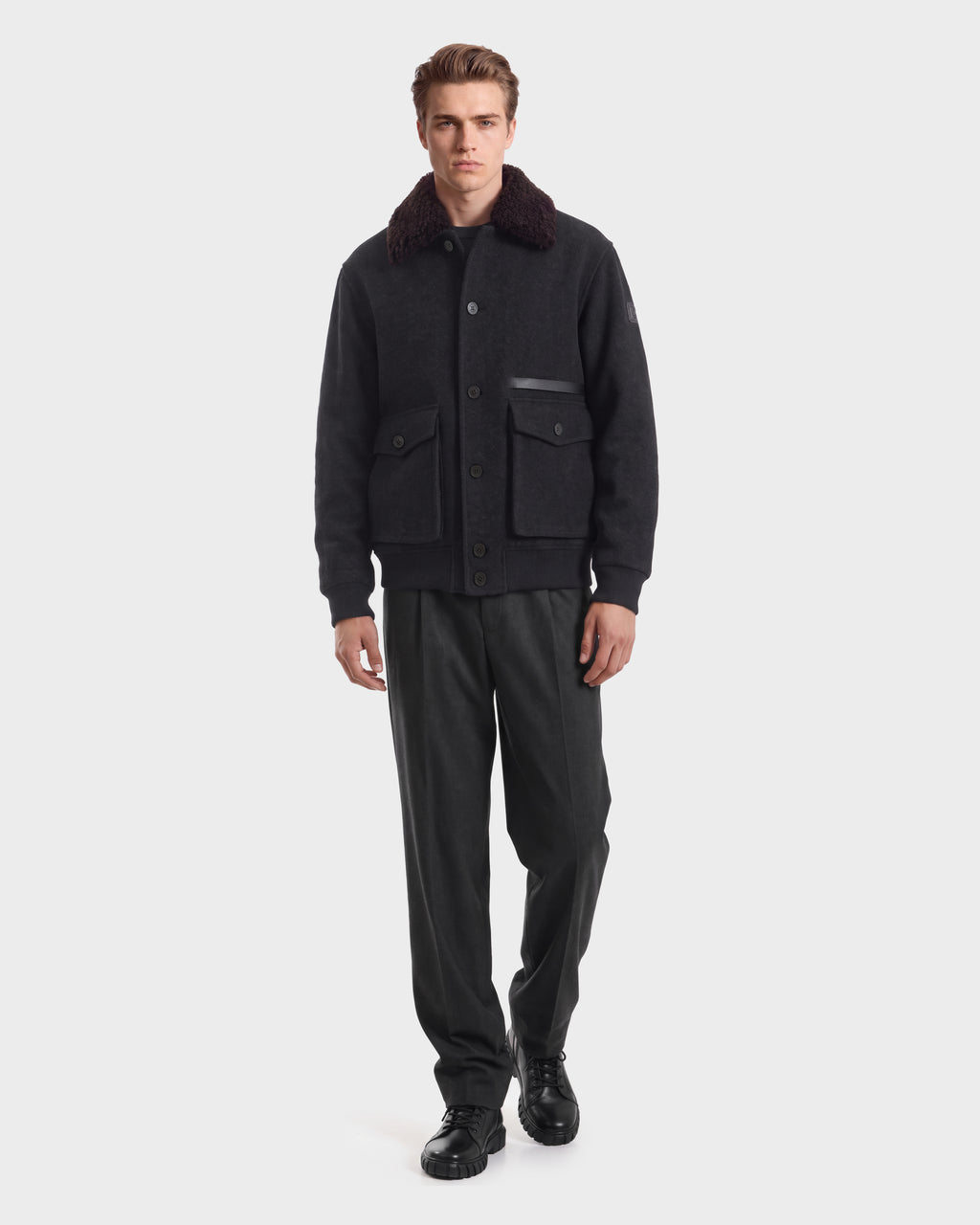 A man wearing a RUDSAK JULIUS - 6126723 Charcoal jacket with a dark faux-fur collar and two front flap pockets, paired with dark trousers and black shoes.