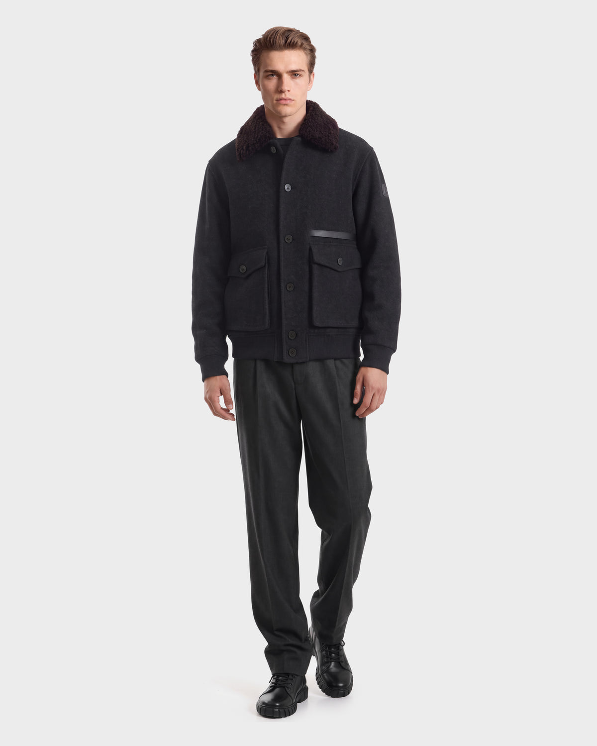 A man wearing a RUDSAK JULIUS - 6126723 Charcoal jacket with a dark faux-fur collar and two front flap pockets, paired with dark trousers and black shoes.