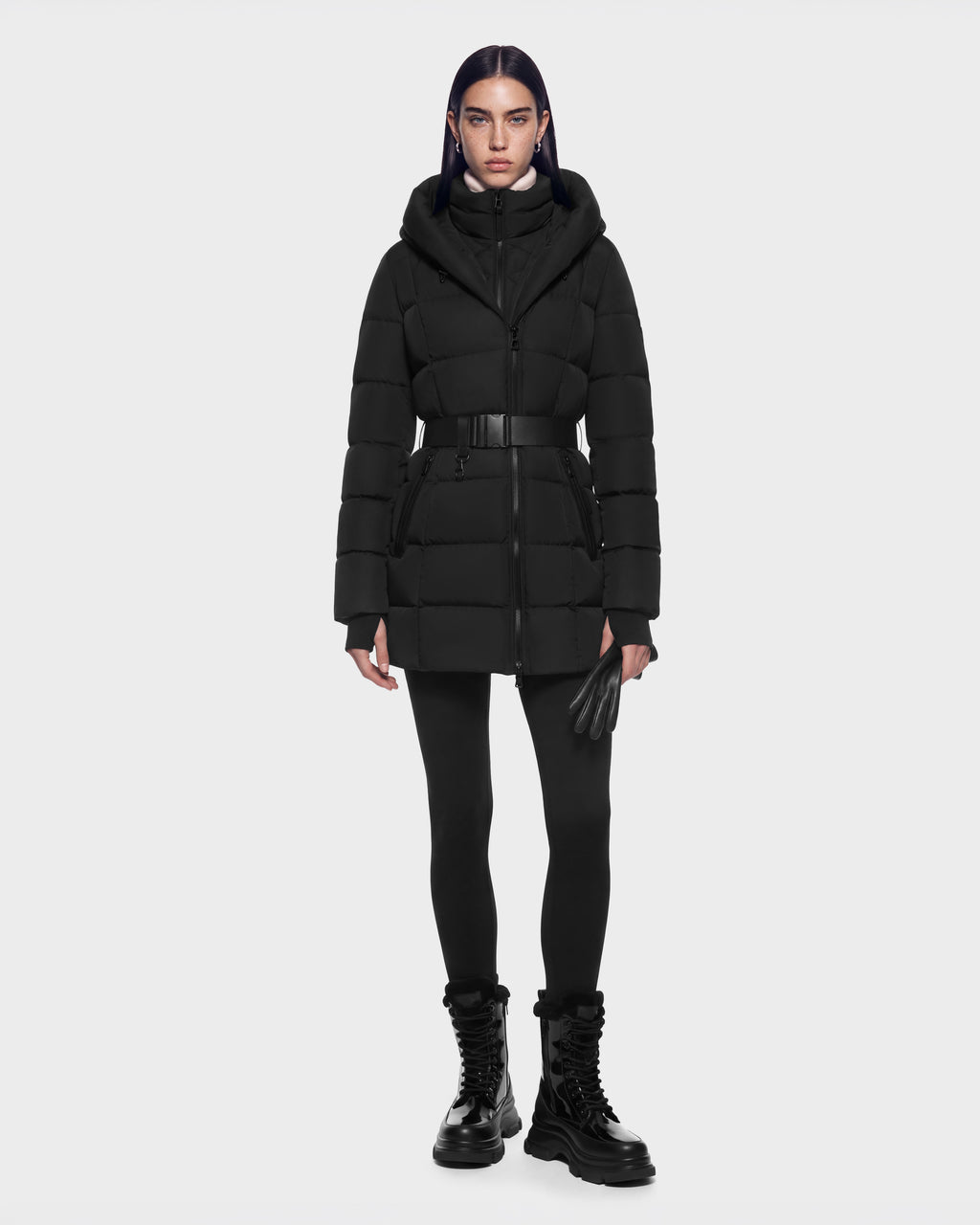 A person wearing a Rudsak TAYLOR M - 8125965 Black belted winter jacket with a hood, black leggings, leather gloves, and black lace-up boots.