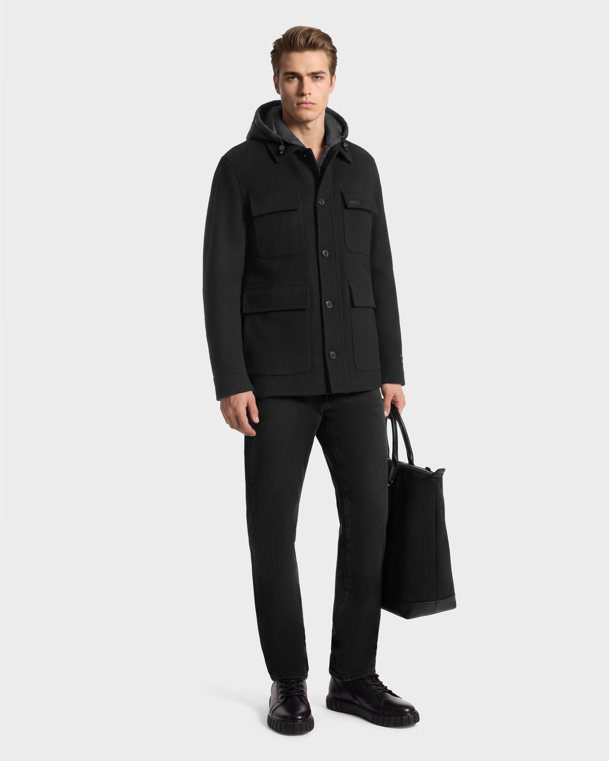 A man wearing a BRUNO - 6126716 Charcoal hooded jacket with four front pockets, black jeans, and black boots, holding a black tote bag.