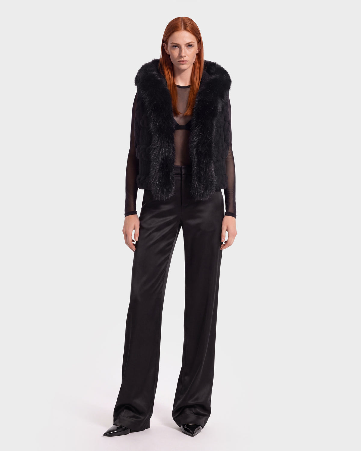 A woman with long red hair models the Rudsak CICELY - 8526686 Black faux fur vest, featuring a faux fur-trimmed hood, layered over a sheer black top and wide-leg black pants against a plain white background—a perfect example of chic winter styling.