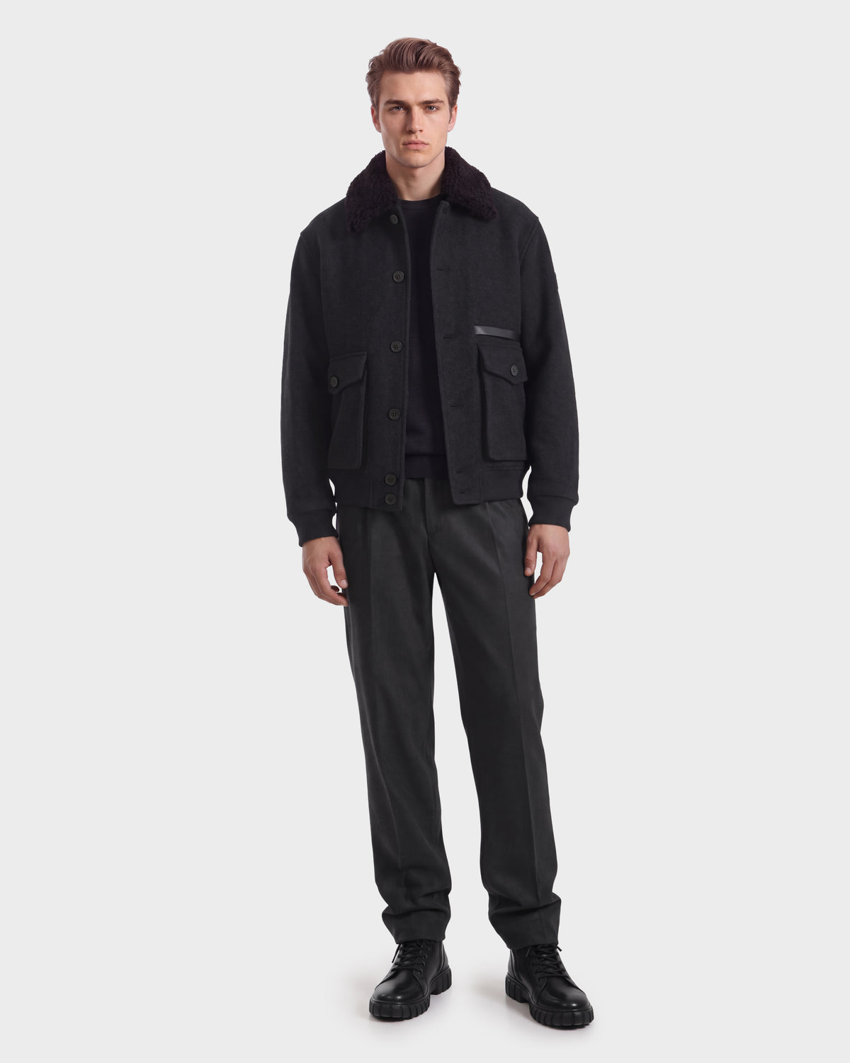 A man wearing a Rudsak JULIUS - 6126723 Charcoal wool jacket with a dark shearling collar, a dark sweater, charcoal trousers, and black leather boots.