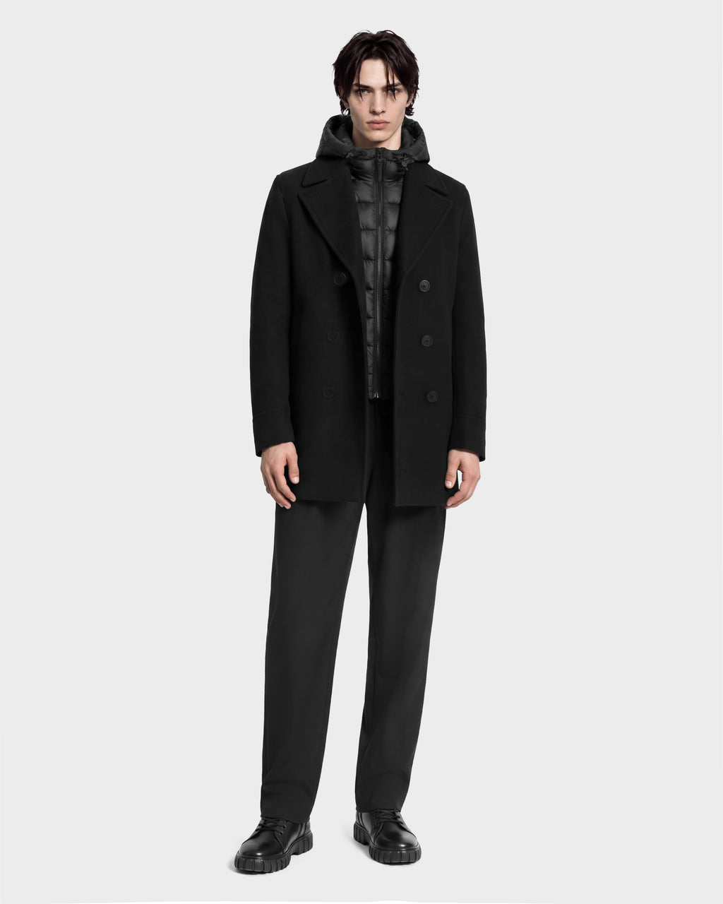 A man wears a black Rudsak LUCIEN - 6126508 Black wool-blend pea coat layered over a black hooded puffer vest with duck down insulation, paired with black trousers and black lug-sole shoes.