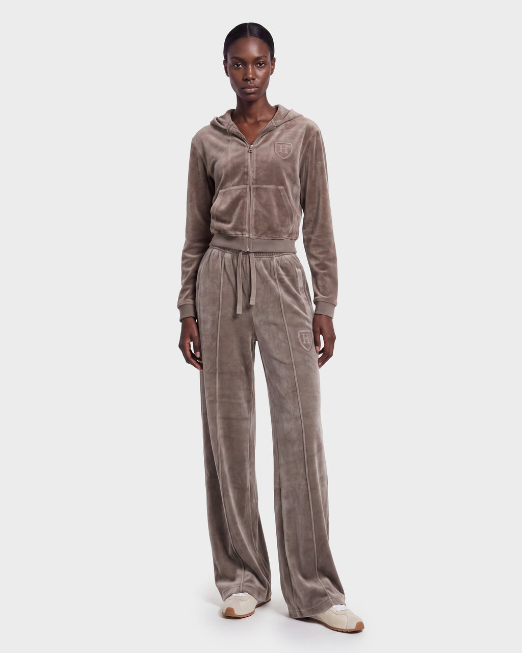 A woman stands against a plain white background, wearing the Rudsak TYRA HV - H526904 Taupe velour tracksuit featuring a zip-up hoodie and wide-leg pants, paired with cream sneakers.