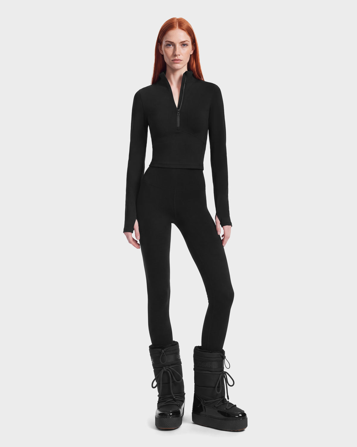 A person wearing a black Rudsak ANITA - 8526579 Black performance-inspired quarter-zip top with a cropped silhouette, paired with black leggings and black winter boots with thick soles and lace-up fronts.