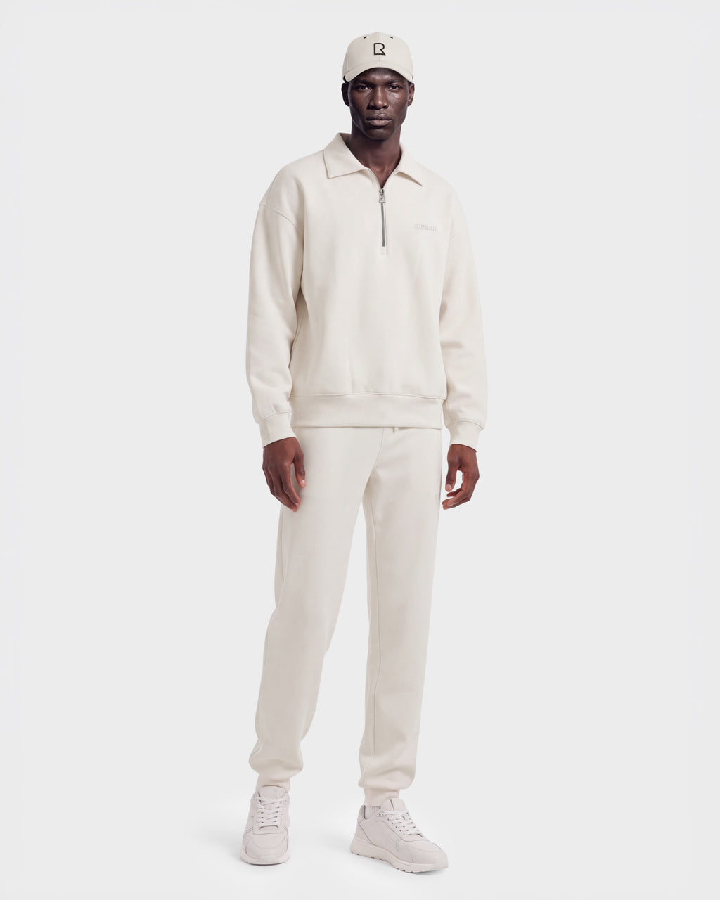 A man wears the IDRIS - 6526500 Vanilla in a cream-colored quarter-zip polo sweater and matching sweatpants, paired with a baseball cap and sneakers for a relaxed silhouette.