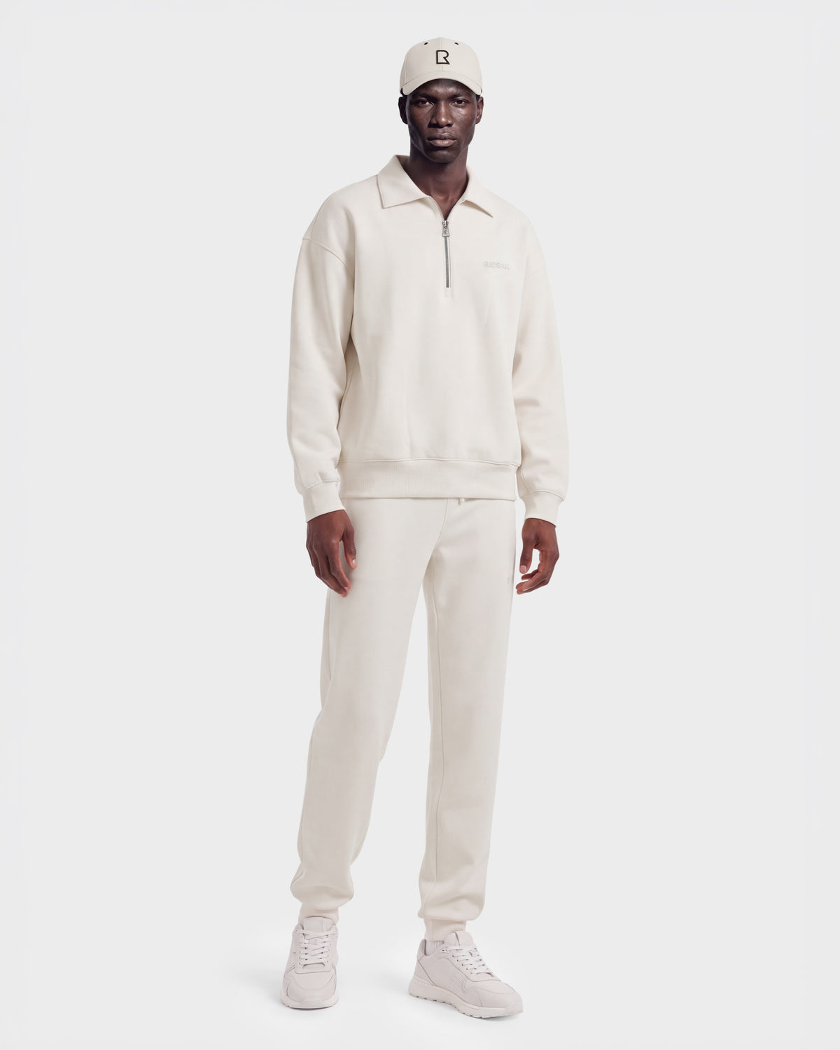 A man wears the IDRIS - 6526500 Vanilla in a cream-colored quarter-zip polo sweater and matching sweatpants, paired with a baseball cap and sneakers for a relaxed silhouette.