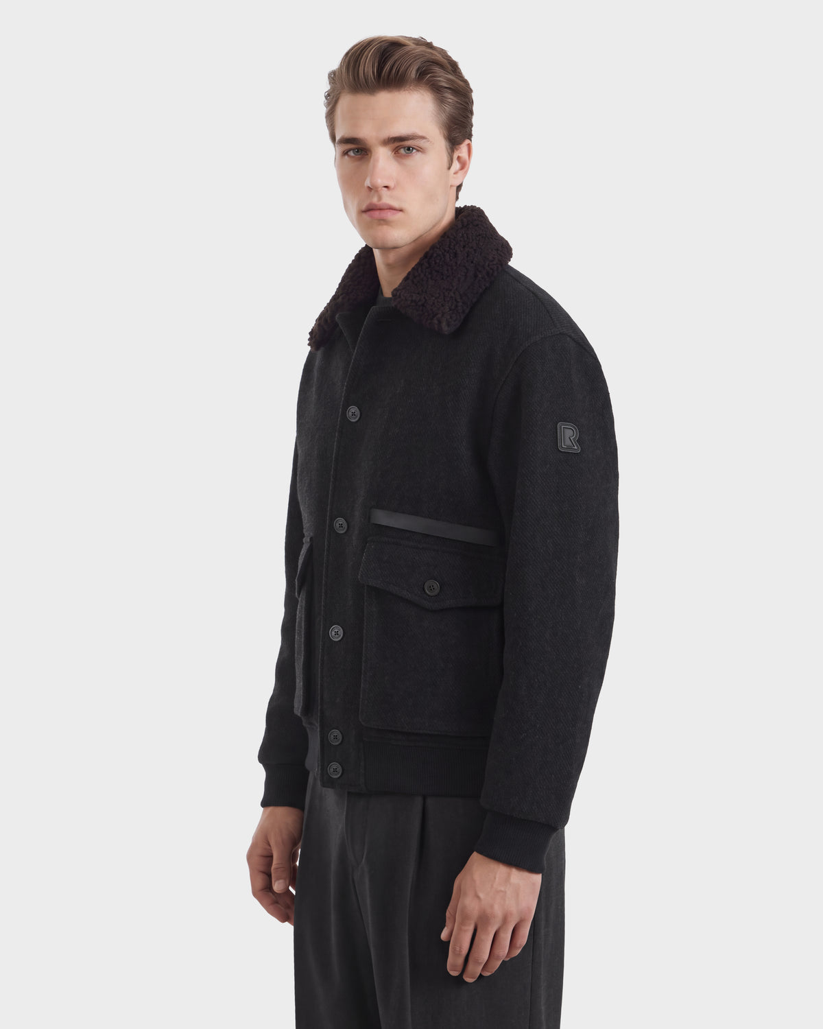 A man wearing a charcoal-colored Rudsak JULIUS - 6126723 bomber jacket with a dark faux-shearling collar and two large front pockets.