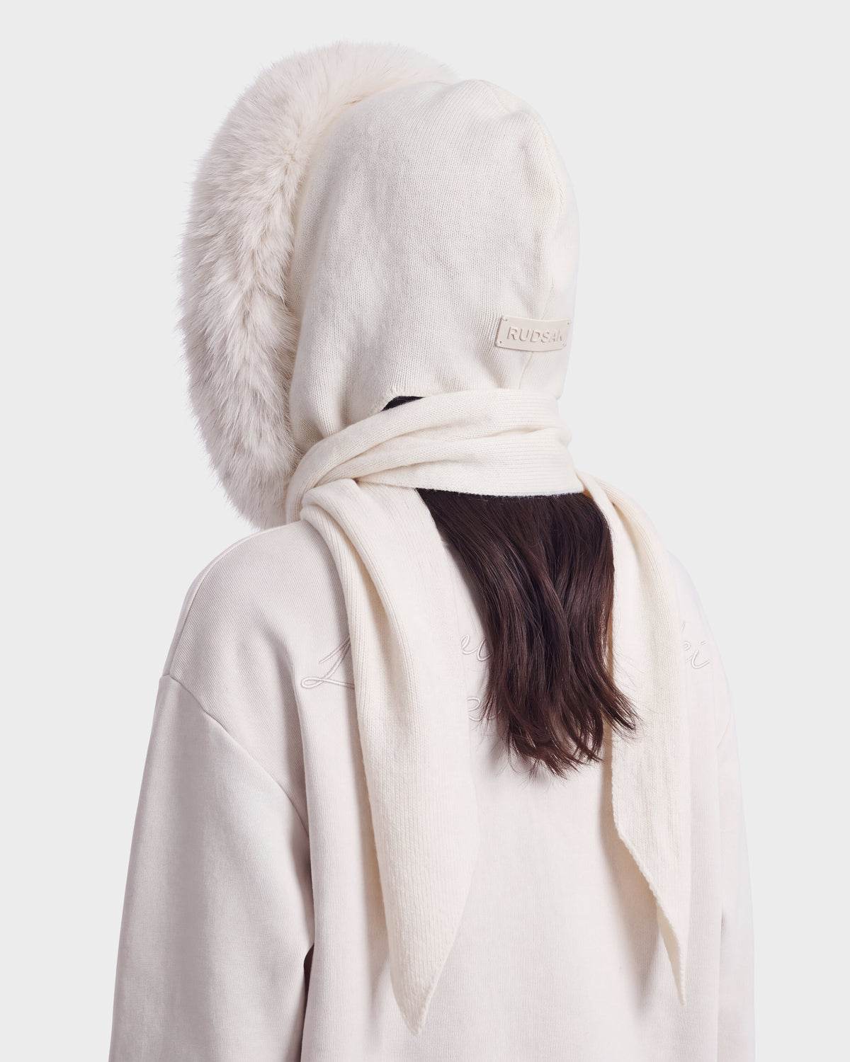 A person is shown from behind, wearing the Rudsak KARYMA - 8426507 Vanilla—a cream-colored hooded coat with a fur trim and a long scarf wrapped around the neck. Only the back of their head and shoulders are visible against a plain background.