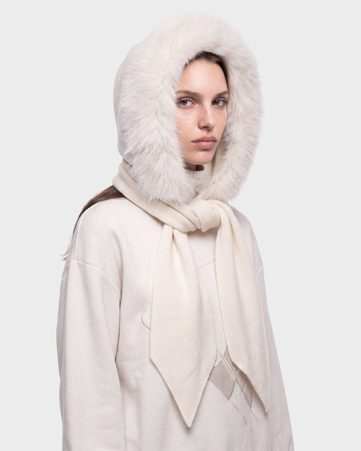 A person wearing the Rudsak KARYMA - 8426507 Vanilla cream-colored sweater with a matching hood featuring faux fur trim, gazing at the camera with a neutral expression against a light gray background.