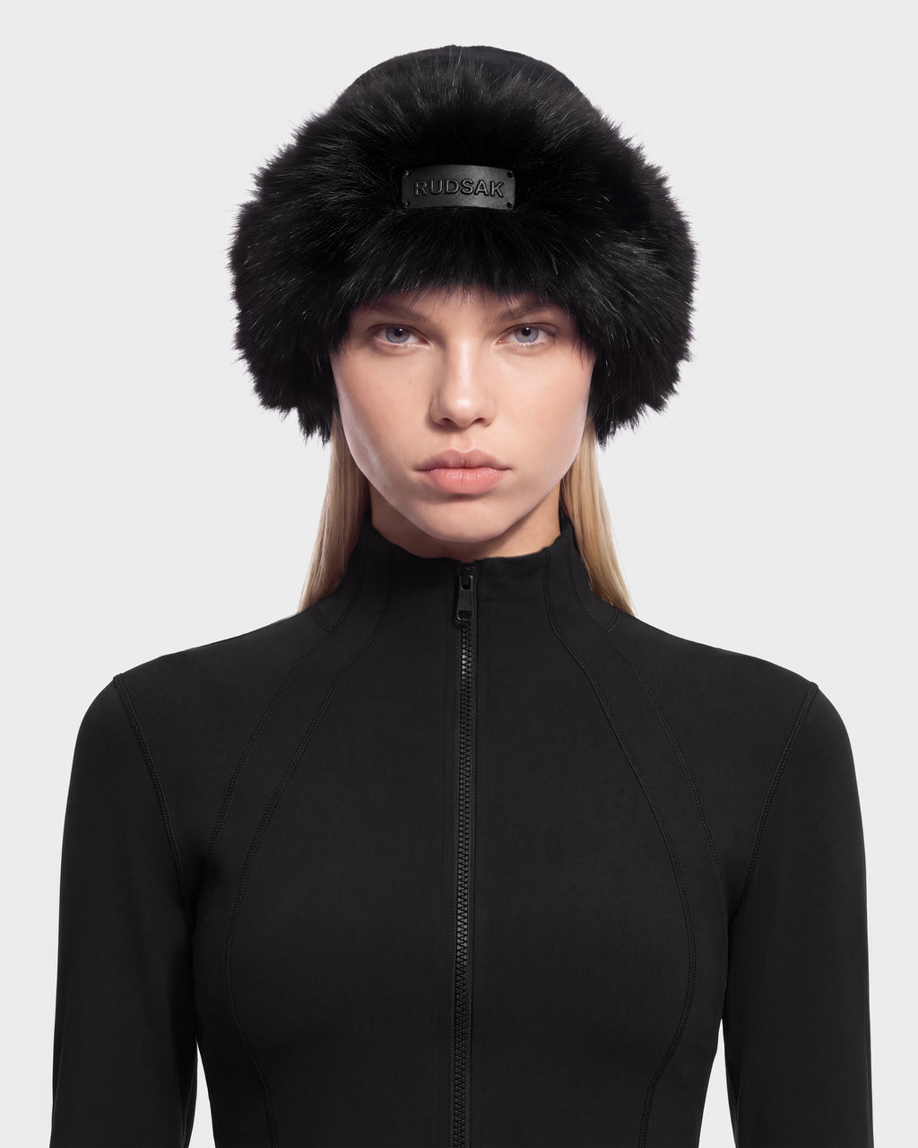 A person wearing a black LORETTA W - 8426510 Black faux fur hat with a branded metal plaque on the front and a black zip-up top.