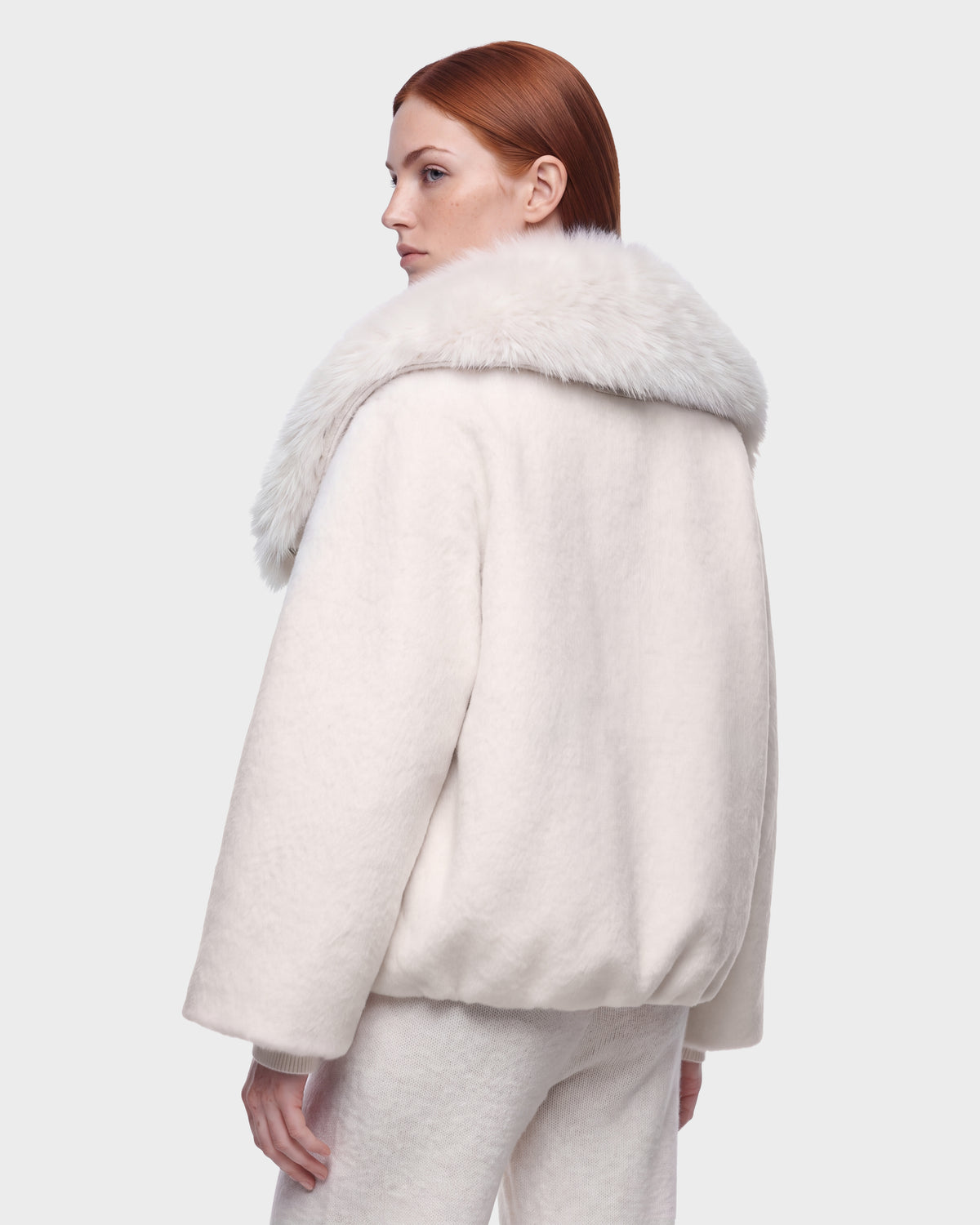 A woman with straight red hair models the Rudsak ZARIA - 8126999 Vanilla puffer, featuring a faux-fur collar and matching white pants, as she looks over her shoulder against a plain light background.