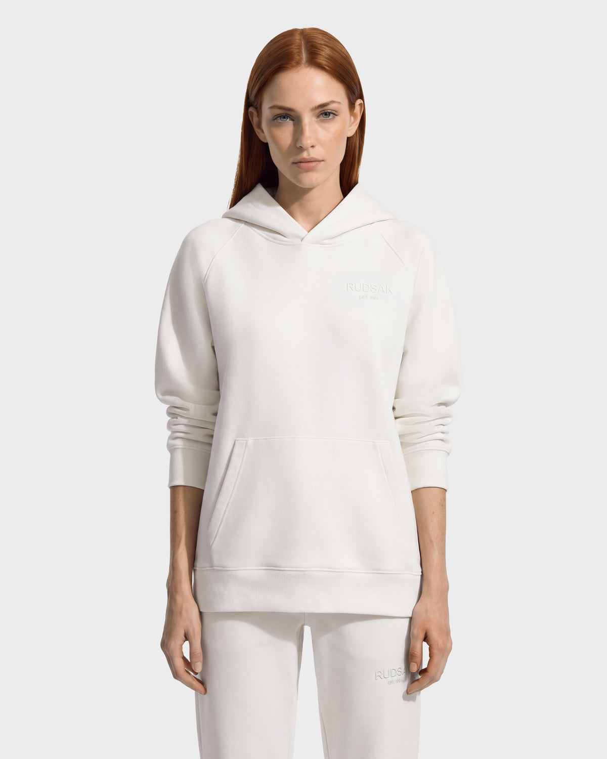 A person wearing a white RUDSAK BRIAR - 9525001 Vanilla W long-sleeved hooded sweatshirt in a premium cotton blend, featuring a front kangaroo pocket and a small logo on the chest.