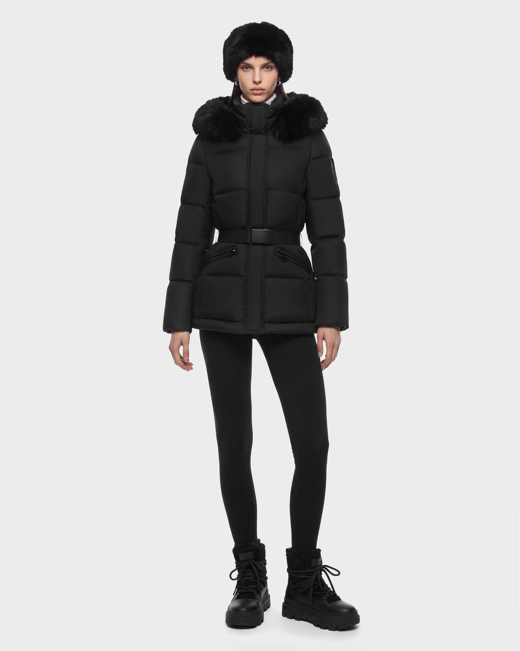 A woman wearing a black Rudsak GRACE FF - 8126535 Black belted puffer jacket with a fur-trimmed hood, a matching fur hat, black leggings, and black lug-sole boots.