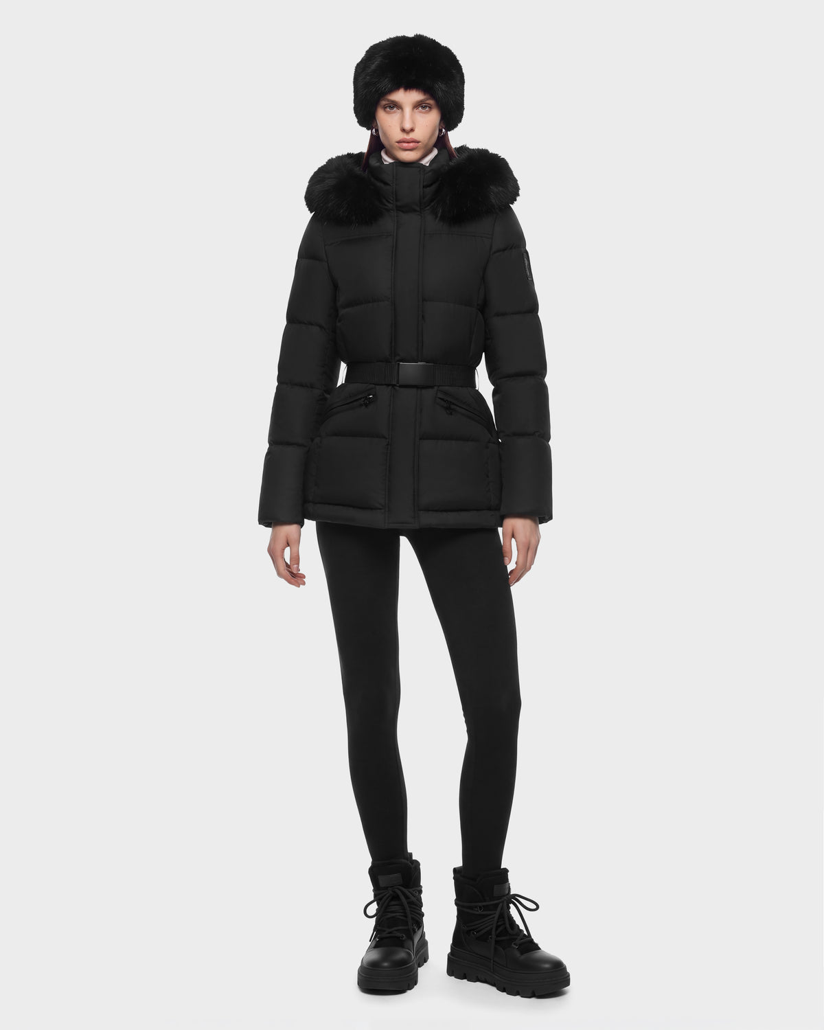 A woman wearing a black Rudsak GRACE FF - 8126535 Black belted puffer jacket with a fur-trimmed hood, a matching fur hat, black leggings, and black lug-sole boots.