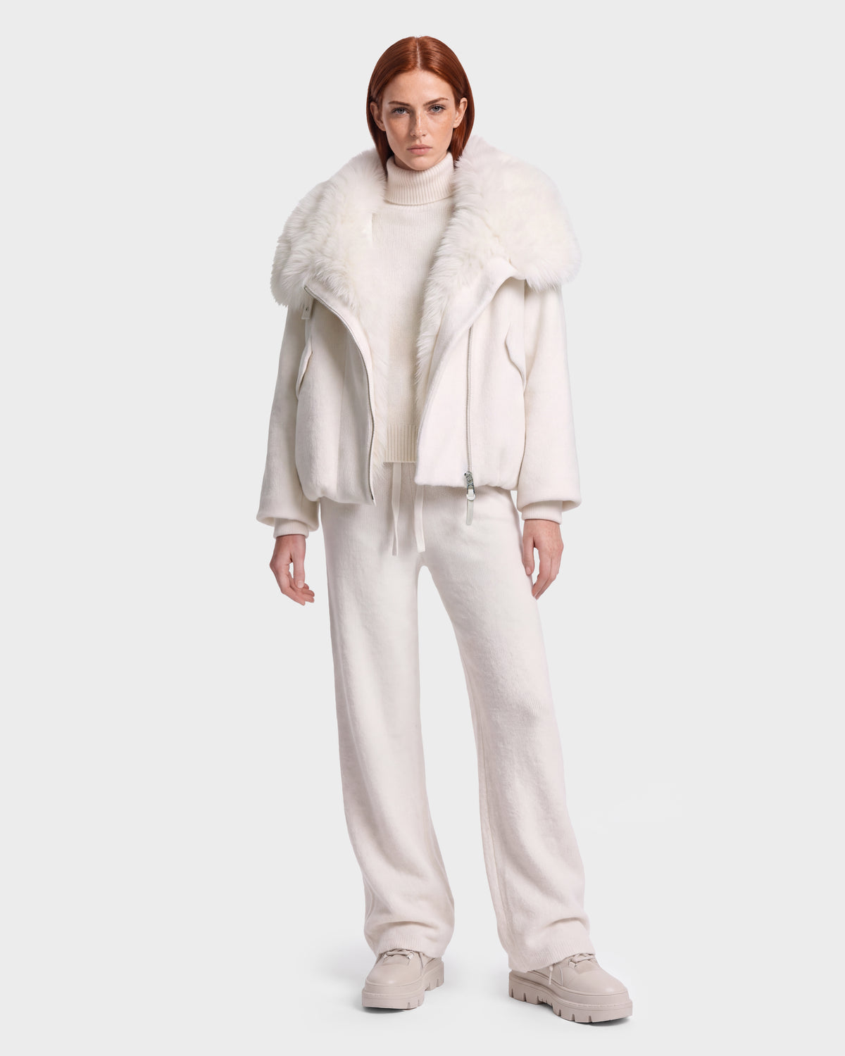 A woman with red hair is seen wearing the Rudsak ZARIA - 8126999 Vanilla cream winter puffer with a faux-fur collar, styled with a turtleneck sweater, drawstring pants, and chunky shoes, standing against a plain white background.