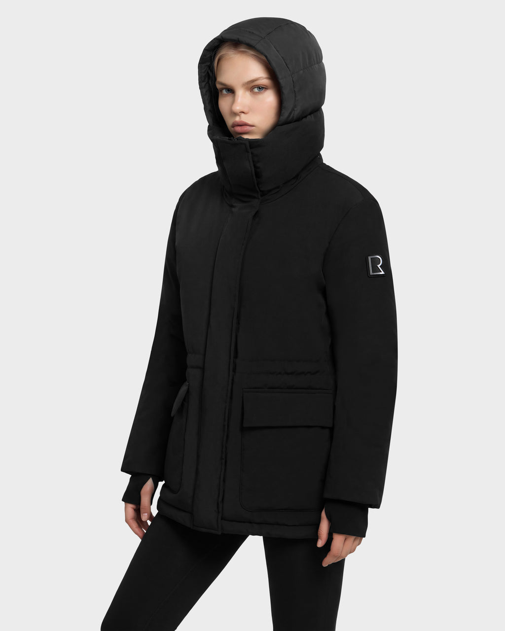 A person wears a black Rudsak BHIMA - 8126515 Black hooded winter parka with a high collar and a logo patch on the sleeve.