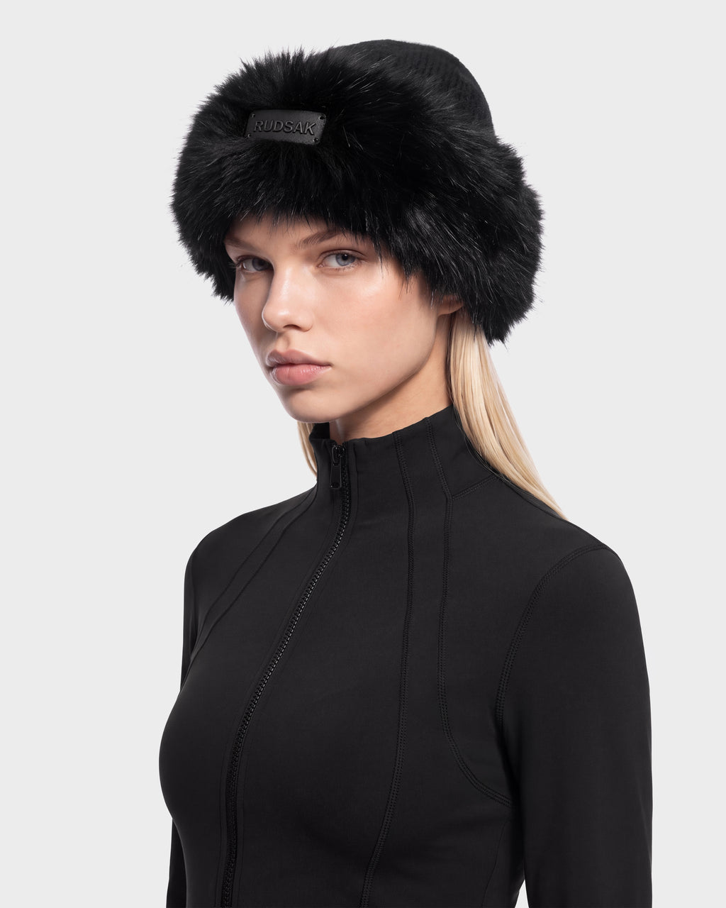 A person wearing a black LORETTA W - 8426510 Black faux-fur trimmed winter hat with a branded plaque on the front and a black zip-up jacket.
