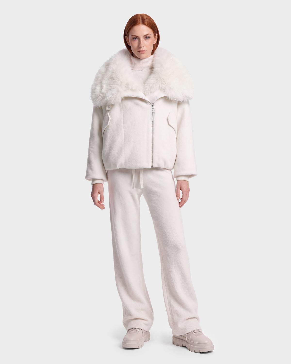 A woman with straight red hair wears the Rudsak ZARIA - 8126999 Vanilla puffer jacket, featuring a plush faux-fur collar, paired with an all-white outfit that includes a turtleneck sweater, loose pants, and chunky shoes, standing against a plain light background.
