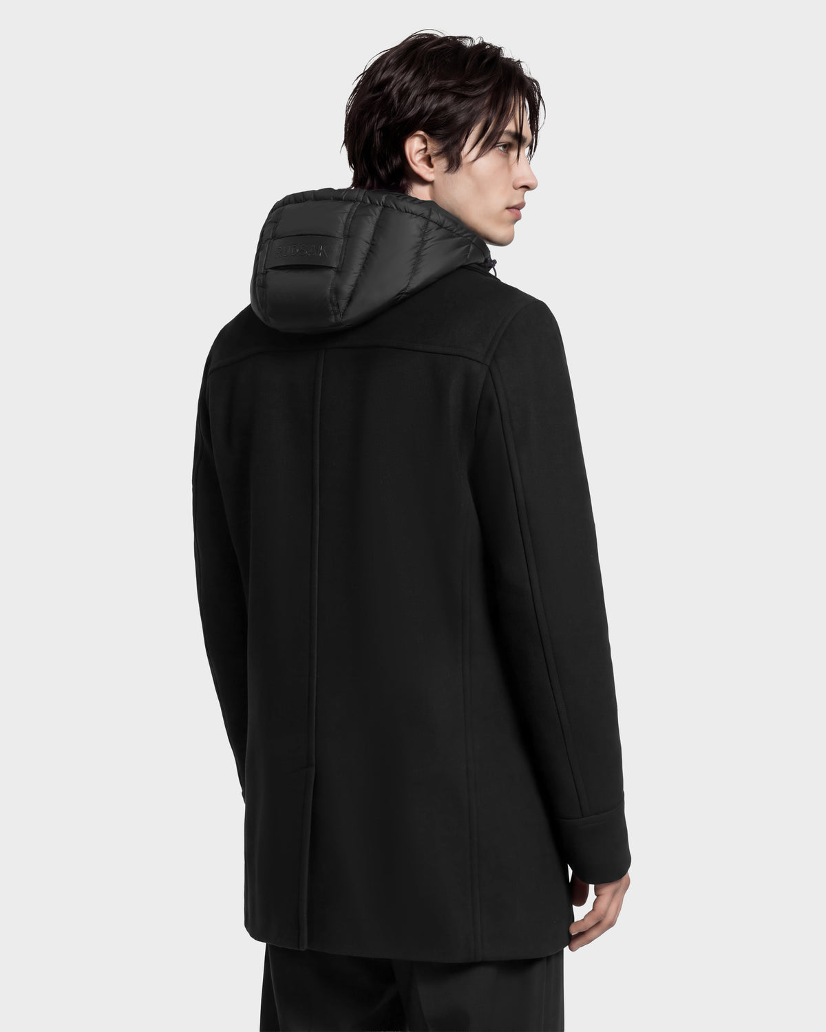 A person seen from the back wearing a black, mid-length Rudsak LUCIEN - 6126508 wool-blend jacket with a puffy hood.