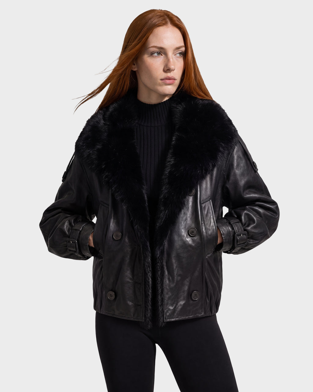 A woman wears a black JANELLE - 8126936 Black leather bomber jacket with a large faux fur collar and button closures, her hands tucked into the pockets.