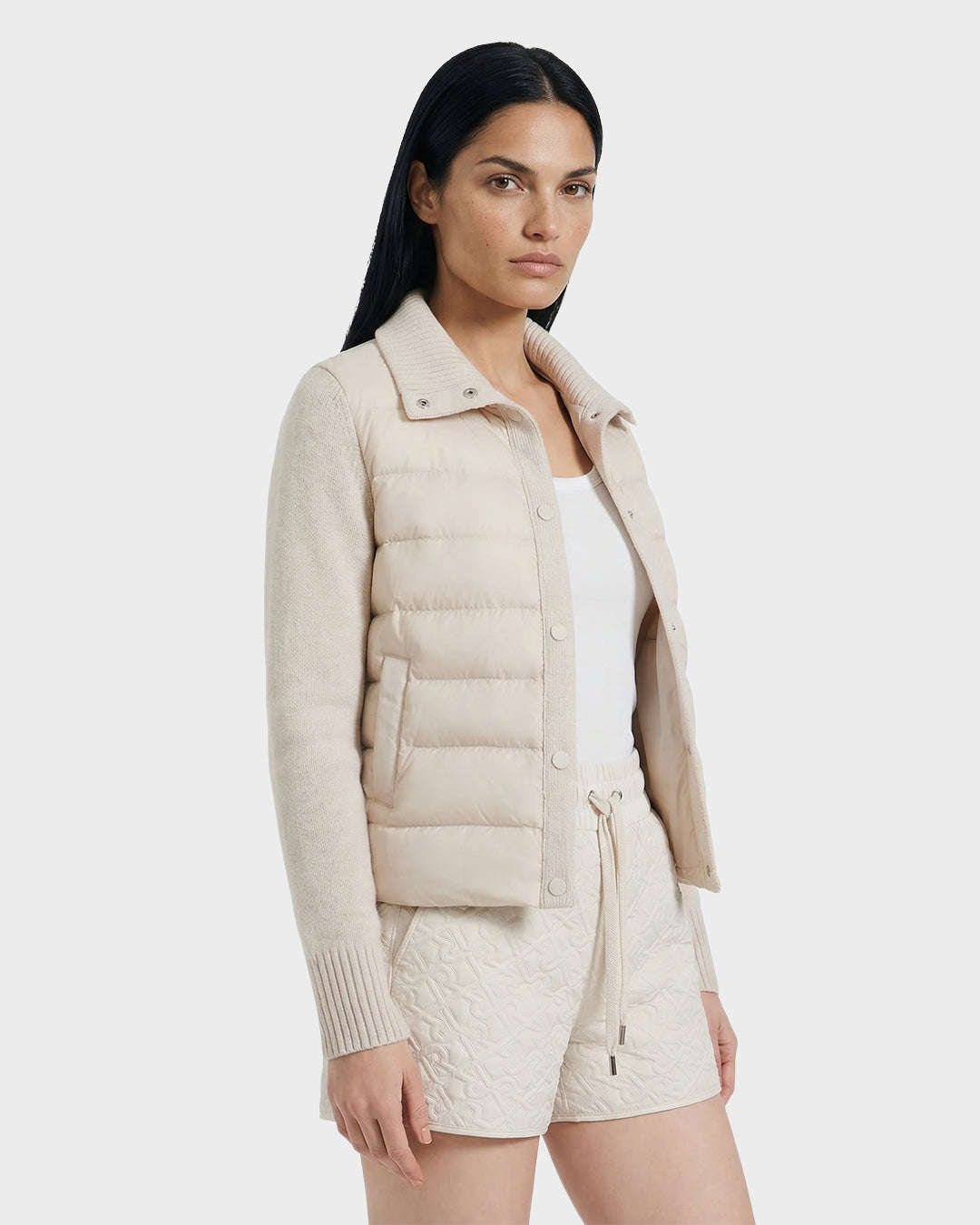A woman confidently poses against a plain light background, wearing the Rudsak ELLIE - 8125635 Vanilla hybrid down jacket over a white top and matching quilted shorts.