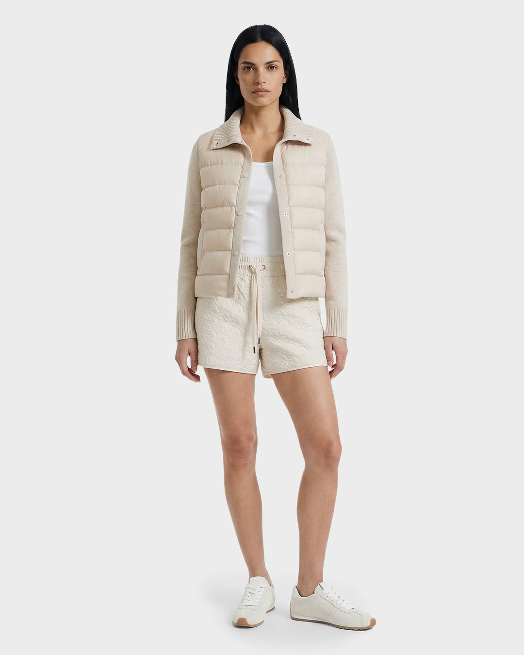 A woman stands facing forward wearing a Rudsak ELLIE - 8125635 Vanilla quilted down jacket over a white top, beige drawstring shorts, and beige sneakers against a plain white background.