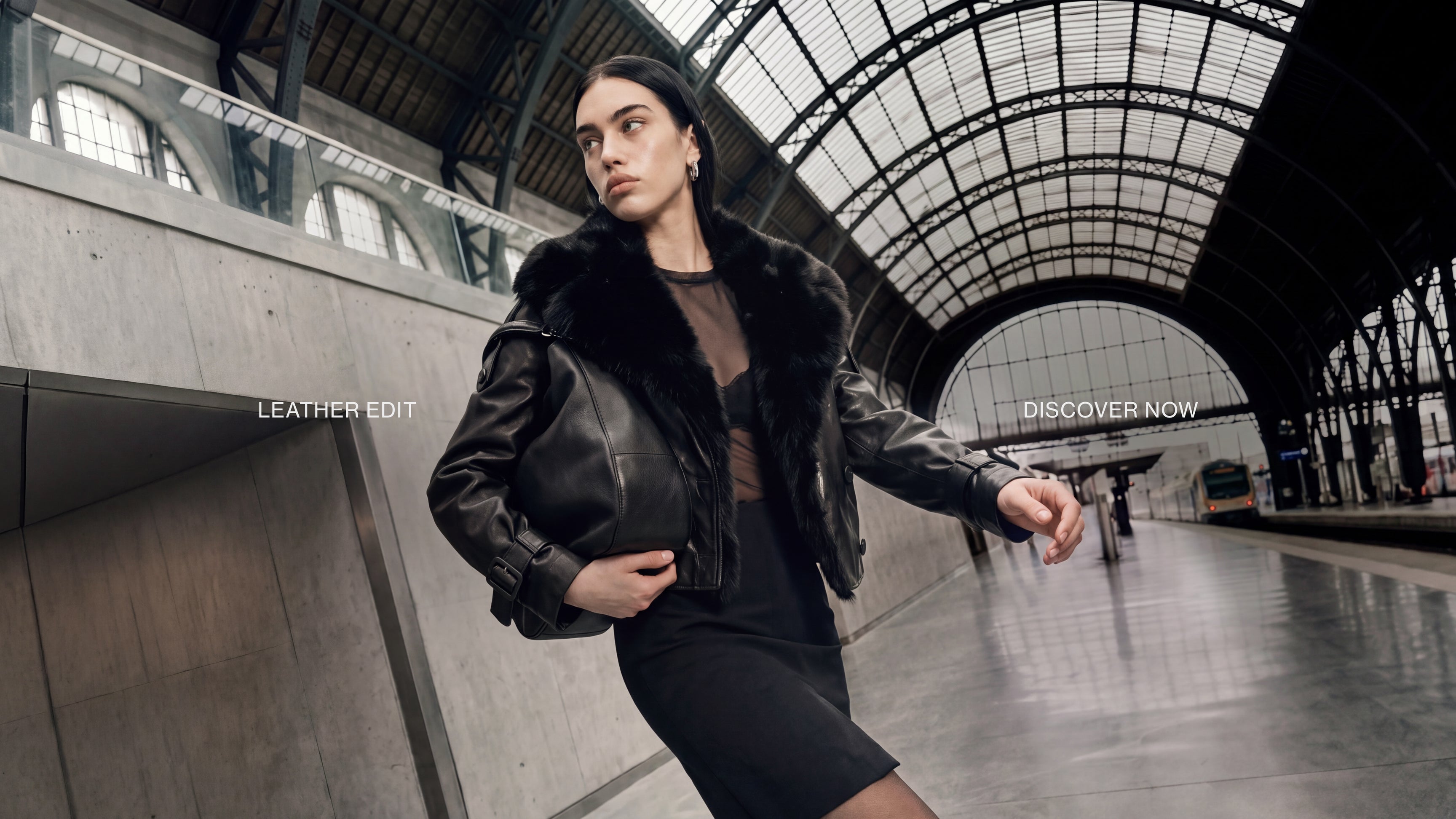 A woman in a black leather jacket with a black fur collar stands in a large, arched train station hall, holding a black leather handbag. 