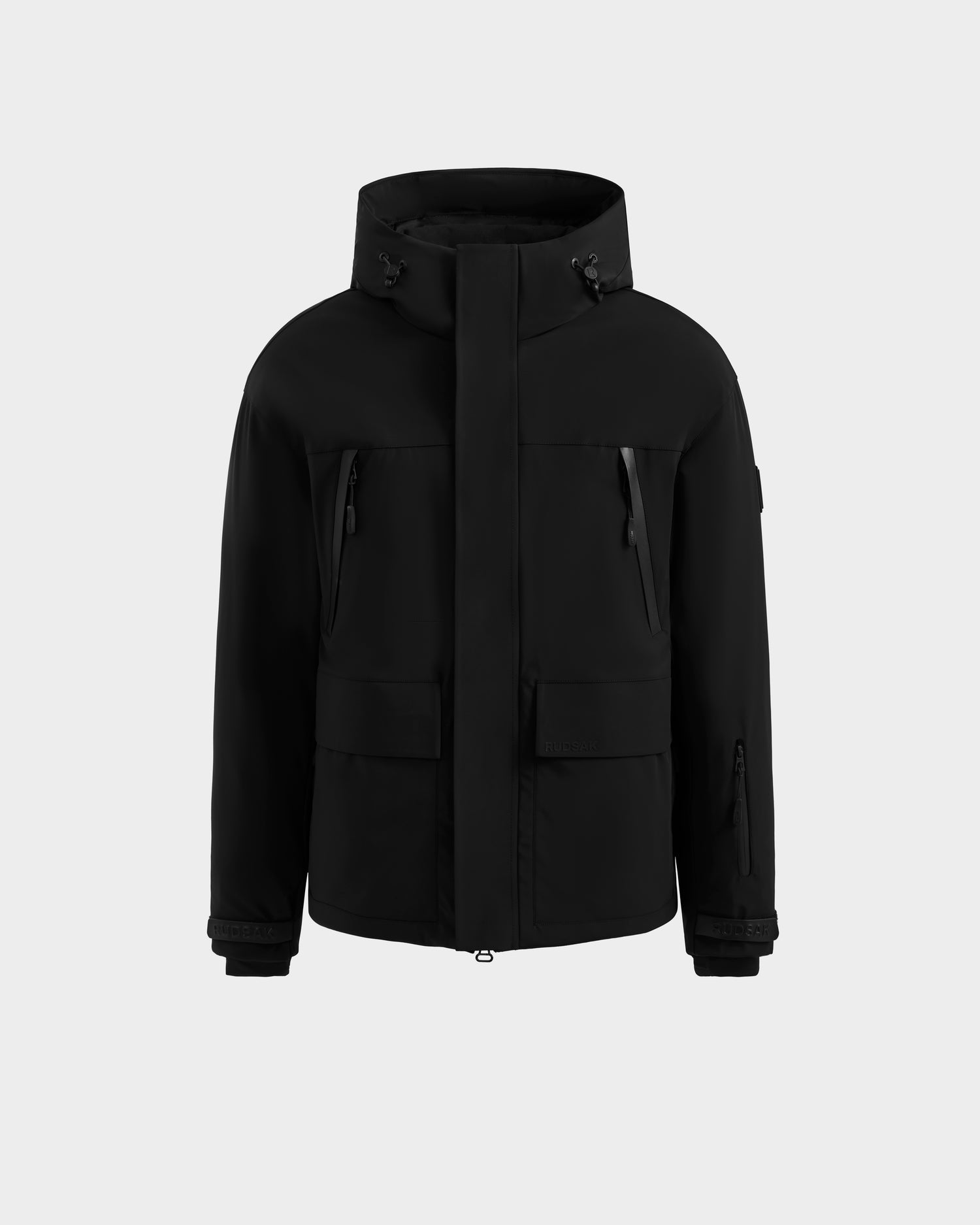 The Rudsak ETHAN - 6125589 Black men’s ski jacket features a front zipper, snap closures, two zippered chest pockets, two large front pockets, and adjustable cuffs, shown against a plain light gray background.